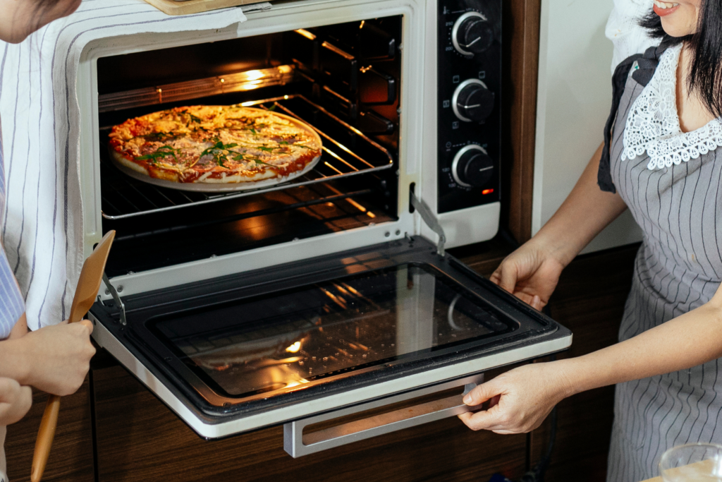 Two people open an oven door showing a cooked pizza inside.