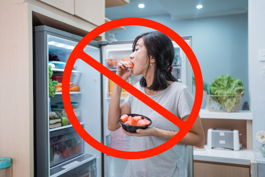A person eats food in front of an opened refrigerator. A red "prohibited" symbol overlays the image, indicating it should be avoided.