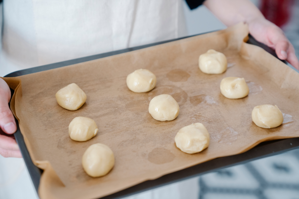 A person holds nine balls of dough sit atop parchment paper on a baking sheet.