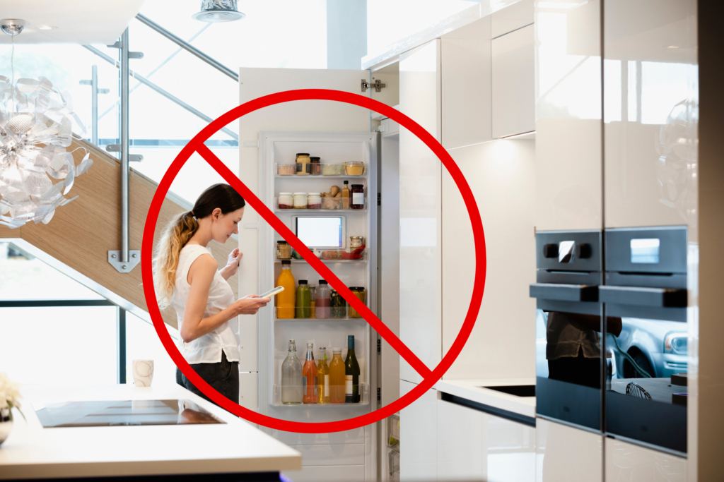 A woman looking at her phone stands in front of an opened refrigerator. A "prohibited" symbol overlays the image, suggesting not to do that.