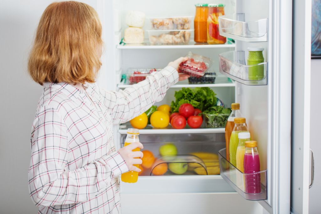A woman holds an open juice bottle and reaches for a pack of fruit inside her fridge.