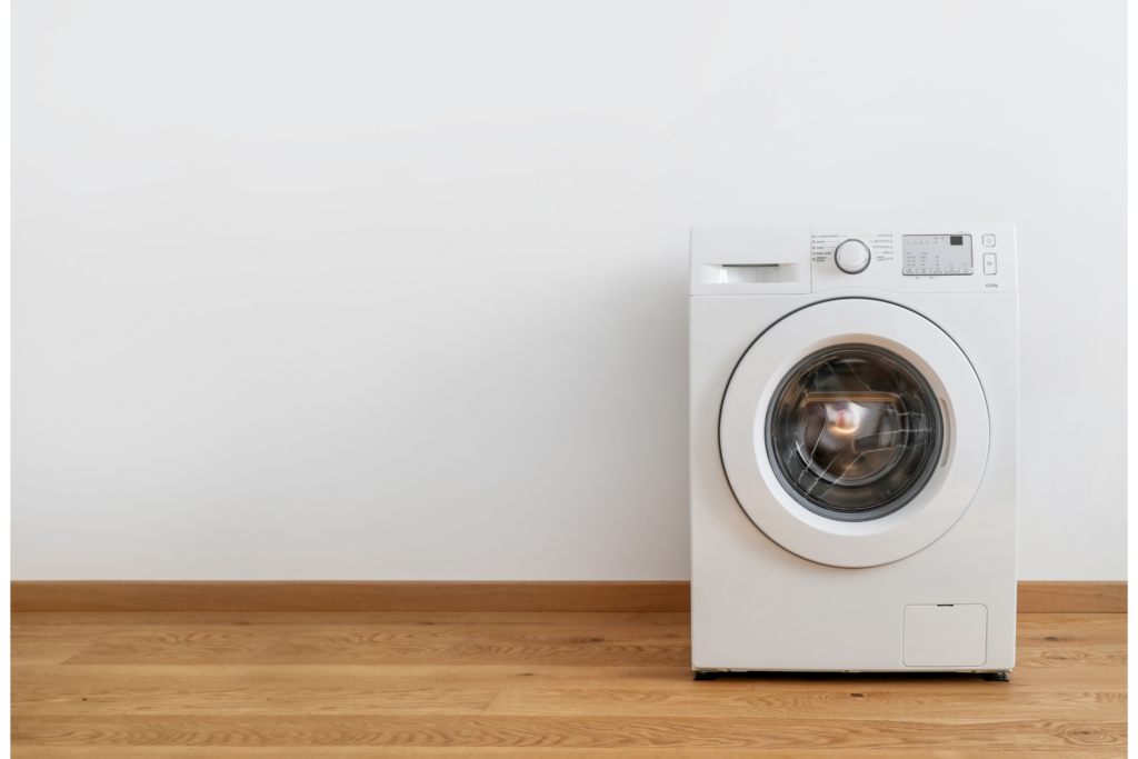 A white front-loading washing machine in front of a white wall with wooden floors beneath.