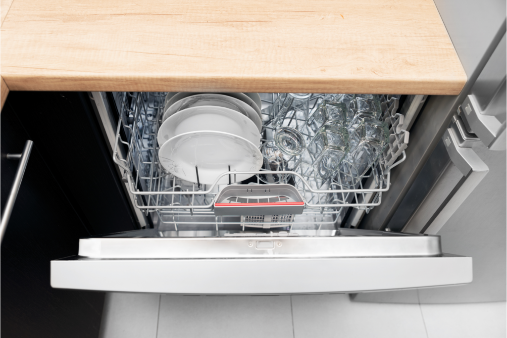 A full dishwasher half open, viewed from above along with a light wooden counter top and silver fridge.